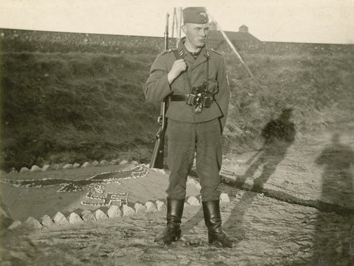 Een op wacht staande Luftwaffe-militiar poseert in Noordwijk voor een wal. Naast hem ligt een perkje waarin met twee kleuren zand en stukken steen een adelaar is aangebracht. Het is een van de vele voorbeelden van dergelijke sierstukjes aan de kust.