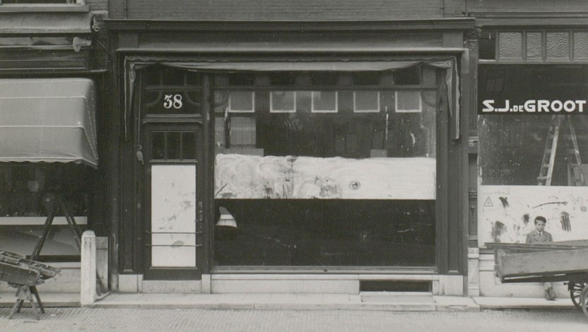 Zwart-wit foto van etalage van winkelpand met nummer 38 op de voordeur. De ramen zijn afgeplakt.